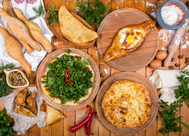 Assorted Georgian pastries rest on wooden boards. Some are topped with fresh greens or cheese, others are filled with egg. Accompaniments include fresh herbs, red chili peppers, and a bowl of flour.