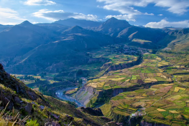 Terraced farmland extends across a valley, with patches of different crops. Mountains rise in the background under a partially cloudy blue sky, creating a picturesque landscape scene.