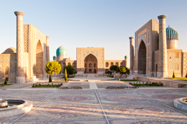A historic square features three grand, ornate buildings with blue domes and intricate arches, surrounding a courtyard with trees and a fountain, under a clear sky at sunset.