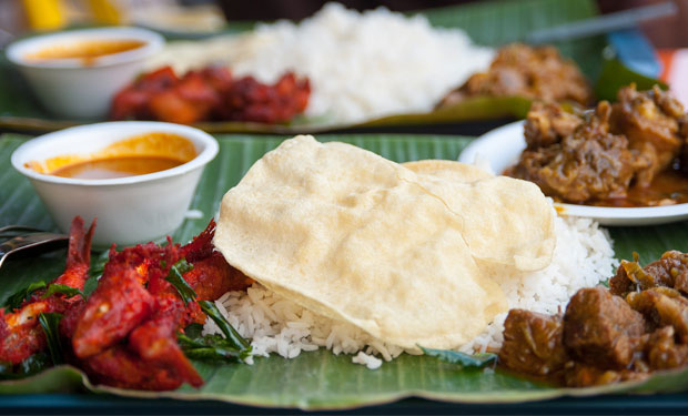Rice served on a banana leaf topped with crispy papadam, accompanied by colorful curries and side dishes in a communal dining setting. Plates and bowls of vibrant sauces surround the dishes.