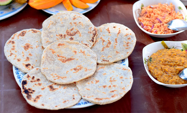 Flatbreads rest on a patterned plate, surrounded by bowls of colorful salads and dips on a dark table, accompanied by sliced mangoes in a dining setting.