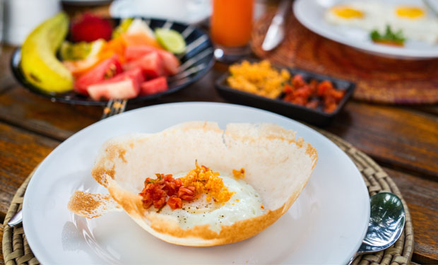 A bowl-shaped hopper with a cooked egg and garnishes sits on a white plate; nearby, fruit, toast, and juice create a vibrant breakfast scene on a wooden table.