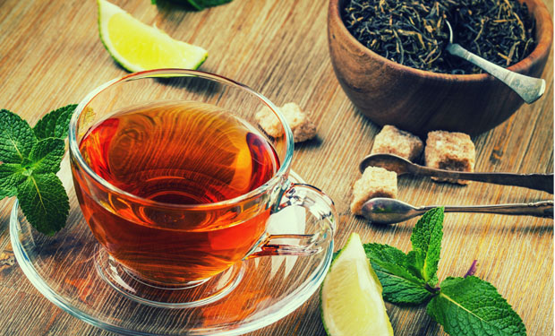 A glass cup holds dark tea, sitting on a wooden table. Surrounding objects include mint leaves, lime wedges, a wooden bowl of tea leaves, sugar cubes, and small silver spoons.