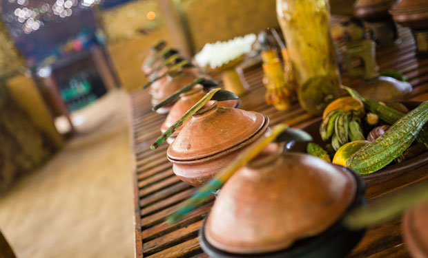 Clay pots with lids and spoons rest on a wooden table, likely containing food. In a rustic setting, various gourds and bottles enhance the earthy ambiance.