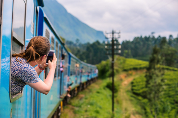 A person holds a smartphone while leaning out a blue train window, capturing a scenic view of lush greenery and distant mountains under a cloudy sky.