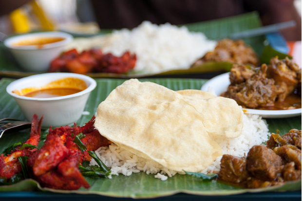 A meal on a banana leaf displays rice, crispy papadum, and various curries, with small dishes of sauce beside it, suggesting a traditional dining setting.