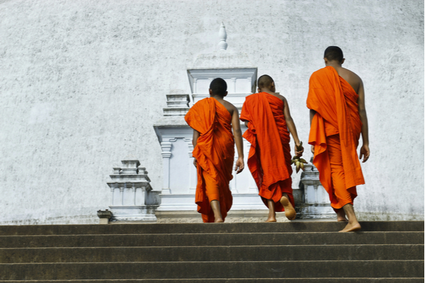 Three monks wearing orange robes ascend stone steps toward a large, white stupa. The stupa's architectural details and the serene setting suggest a place of worship or meditation.