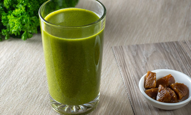 A glass filled with a green smoothie sits on a textured surface, accompanied by a small white bowl containing chunks of brown solid food, with greenery in the background.
