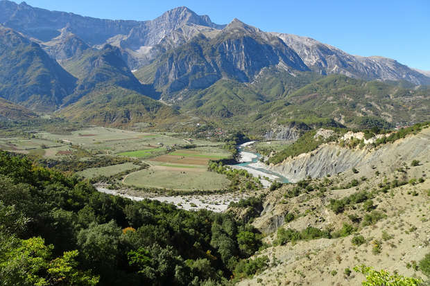 A river winds through a lush valley surrounded by rugged mountains under a clear blue sky. Verdant fields and patches of forest enhance the landscape’s vibrant greenery.