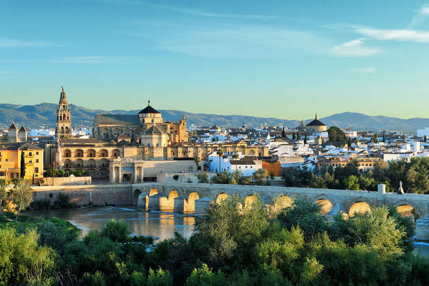 Historic stone bridge spans a river, leading to a city with a large, ornate cathedral and diverse architecture. Rolling hills and mountains form the backdrop under a clear blue sky.