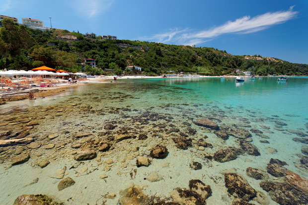 Clear turquoise water reveals a rocky seabed, with nearby sandy beach dotted with sun loungers and umbrellas. People swim and relax, surrounded by lush green hills under a blue sky.