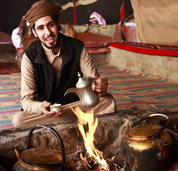 A man, seated cross-legged, pours tea from a metal pot into a small cup over a fire pit. He's in a traditional tent with patterned matting.