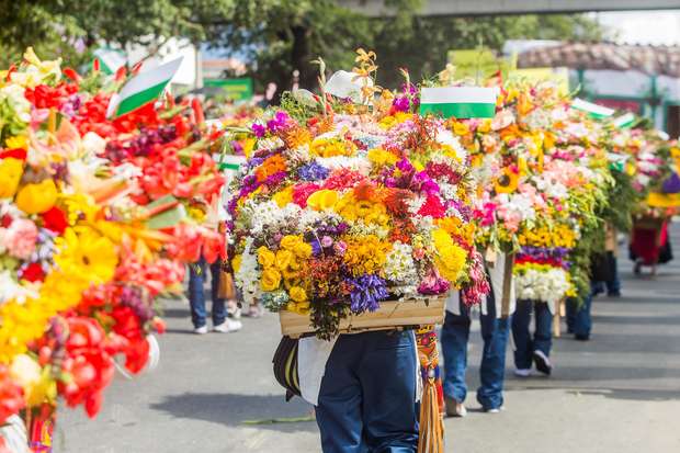 People carry large, colorful flower arrangements on their backs in a parade. Bright blooms include yellow, pink, and red flowers. The event takes place outdoors, amid other participants and spectators.