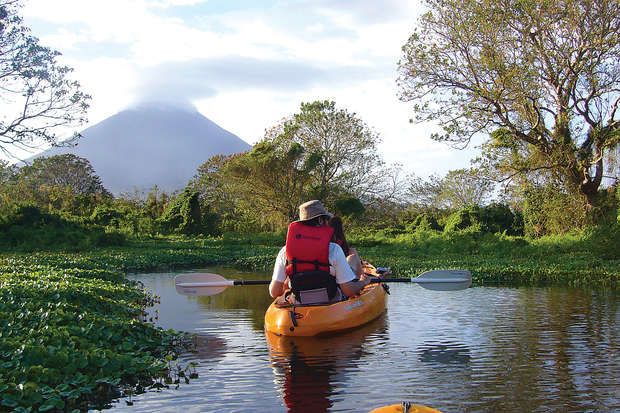 A person paddles an orange kayak through a narrow, calm waterway. The surroundings are lush with greenery, and a distant, cloud-topped mountain is visible against the sky.