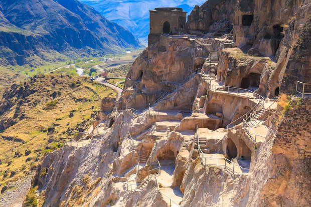 Ancient rock-hewn caves form a historic monastery, cascading down a steep mountainside. Staircases and railings connect the caves. Surrounding landscape features rugged mountains and a distant winding river.