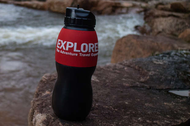 A black water bottle with a red band labeled "EXPLORE: The Adventure Travel Experts" rests on a rock near a flowing river, surrounded by rugged, natural terrain.