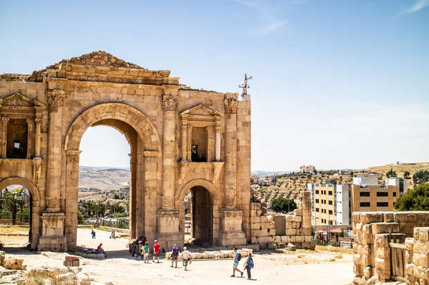 Ancient stone archway stands prominently, with people walking beneath it, surrounded by ruins and modern buildings in the distant background under a bright blue sky.