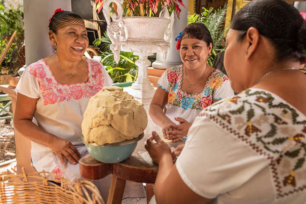 Women in traditional embroidered clothing knead dough at a small wooden table, laughing and engaging with each other. The setting includes lush plants and decorative ceramics in a bright outdoor space.