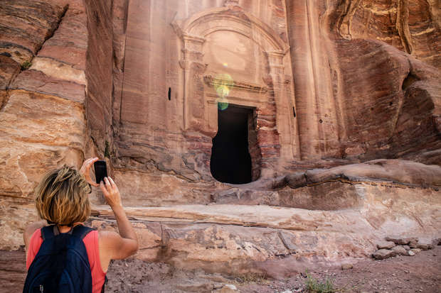 A person photographs a carved rock structure, resembling a tall, ancient architectural façade with a dark entrance, surrounded by rugged, reddish rock formations in a sunny, outdoor setting.