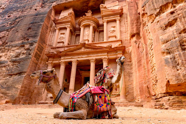 A camel rests adorned with colorful blankets in front of the ancient rock-carved facade of Al-Khazneh, Petra, surrounded by towering sandstone cliffs.