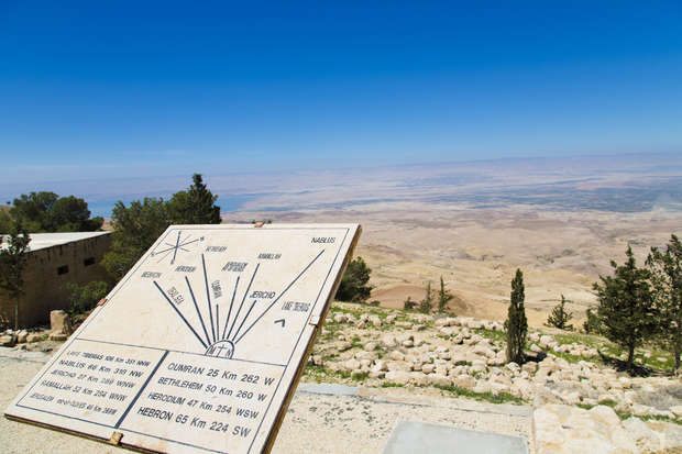 A directional sign points toward various city distances amid a rocky, arid landscape with sparse vegetation and distant hills under a clear blue sky.