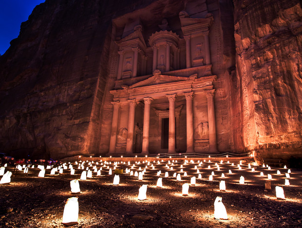 Ancient rock-carved building illuminated at night, surrounded by numerous glowing lanterns placed on the ground, creating an enchanting atmosphere. The architectural facade features columns and intricate carvings.