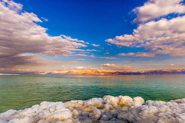 Salt formations line a tranquil, turquoise shore; fluffy clouds scatter across a vibrant blue sky, casting shadows over distant, sunlit hills, creating a serene, picturesque landscape.