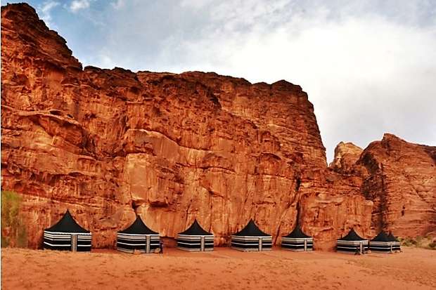 Five black, striped tents stand aligned at the base of a towering, reddish rock cliff, under a partly cloudy sky in a desert landscape.