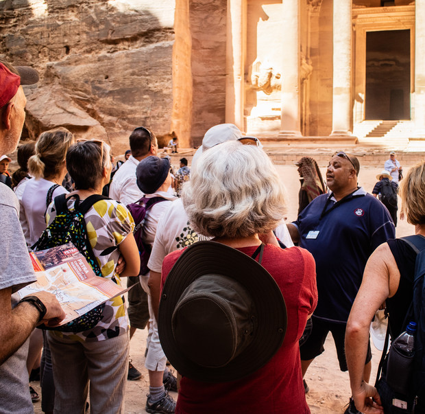 A group of tourists attentively listens to a guide near an ancient stone structure with carved pillars and steep steps in a sunlit, rocky setting.