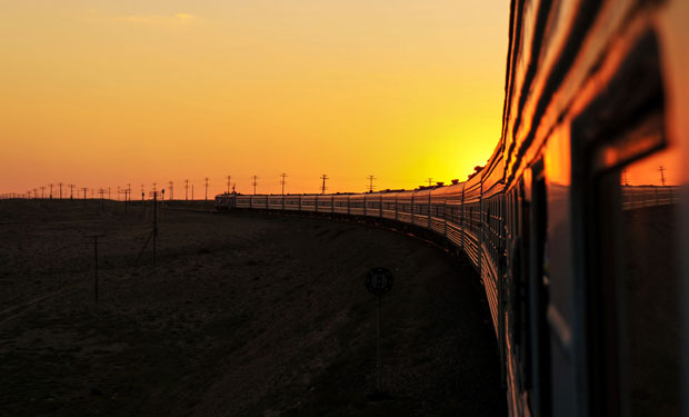 A train travels along curved tracks during sunset, reflecting golden light. The surrounding landscape features open fields and distant power lines against an orange sky.