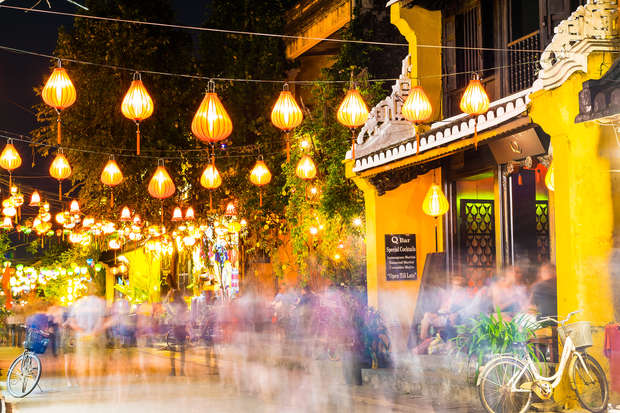 Lanterns hang above a bustling street filled with blurred pedestrians. Vibrant yellow building displays a sign reading: "Q Bar - Spirit Lounge." A parked bicycle rests nearby.