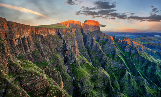 Steep cliffs bathed in warm sunset light rise dramatically, covered in patches of green vegetation. The expansive landscape stretches under a vibrant sky with scattered clouds.