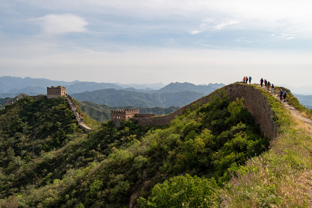 People walk along a section of a historic stone wall atop a lush, green ridge, with watchtowers in the distance and a mountainous landscape under a partly cloudy sky.