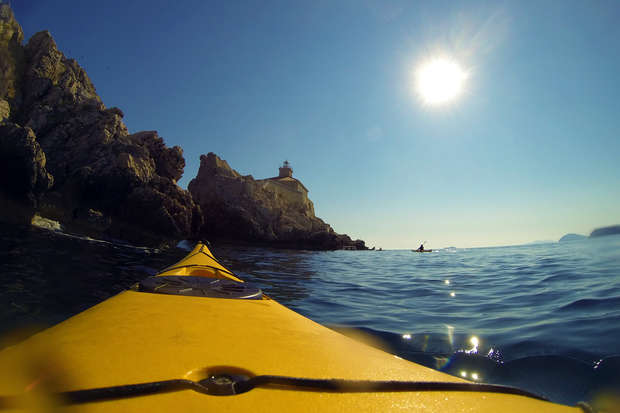 A yellow kayak glides on calm ocean water near rocky cliffs under a bright sun, with a distant small structure atop the rocks and clear blue sky overhead.