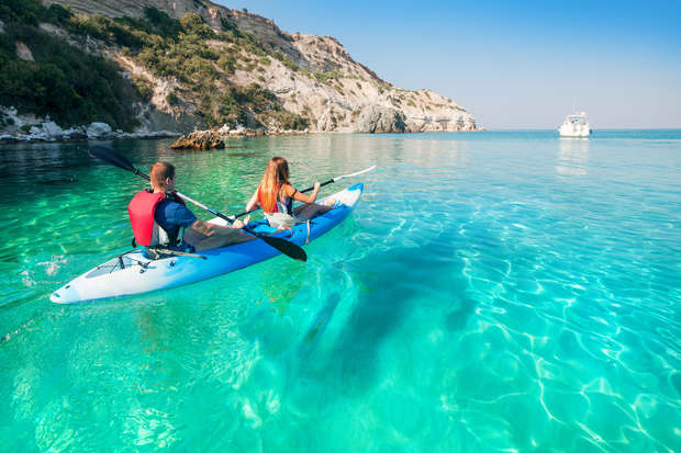 Two people paddle a kayak through clear turquoise water near a rocky coastline, with a distant boat visible under a clear blue sky.