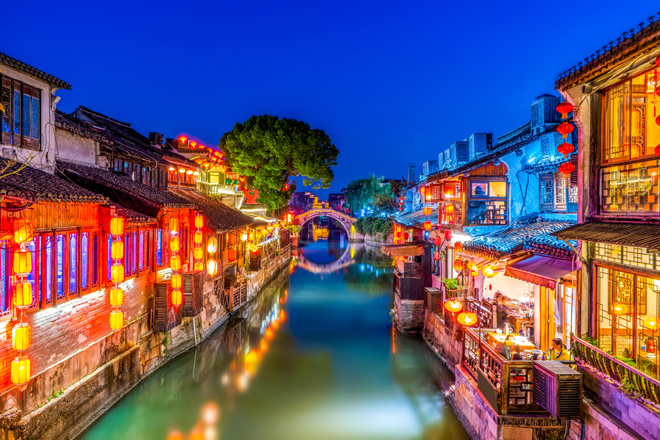 Colorful lanterns illuminate traditional wooden buildings lining a narrow canal, reflecting vibrant hues on the water. An arched stone bridge connects both sides under a clear, deep blue evening sky.