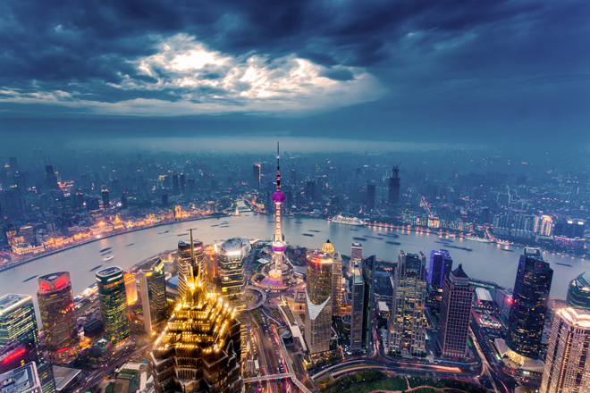 Skyscrapers with vibrant lights cluster around a river, dominated by a tall tower with a purple sphere. The cityscape extends under a dramatic, cloudy evening sky.