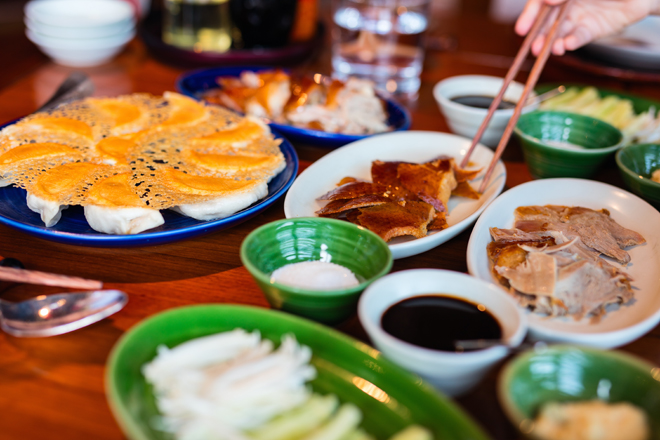 A variety of dishes on a wooden table includes dumplings with crispy golden edges. A hand uses chopsticks to pick crispy skin from a plate. Green bowls hold condiments, surrounding sauce dishes.