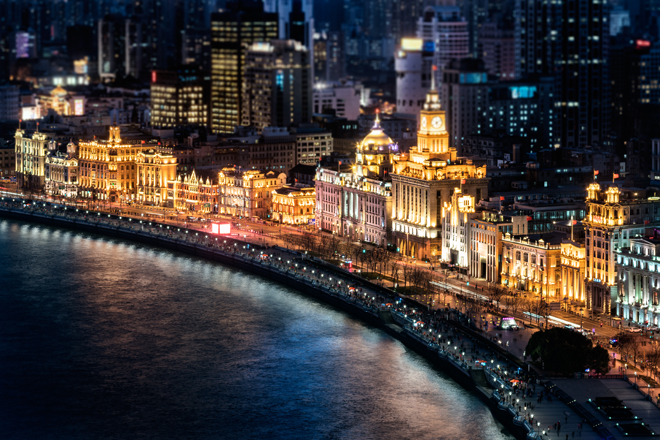 Illuminated historic buildings glow brightly along a waterfront promenade at night. Skyscrapers rise in the background, while reflections shimmer on the water in an urban cityscape setting.