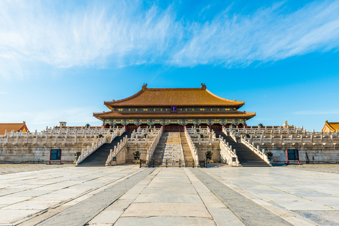An ornate, historic building with a golden roof stands prominently. Stairs lead up to it, surrounded by decorative stone balustrades. The scene is set under a bright blue sky.