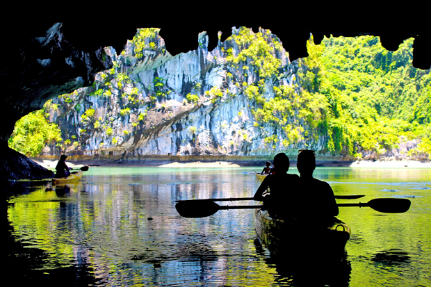 Silhouetted kayakers paddle through a cave, entering a vibrant lagoon surrounded by lush, rocky cliffs. Sunlight highlights the greenery and reflects off the water's surface, creating a serene atmosphere.
