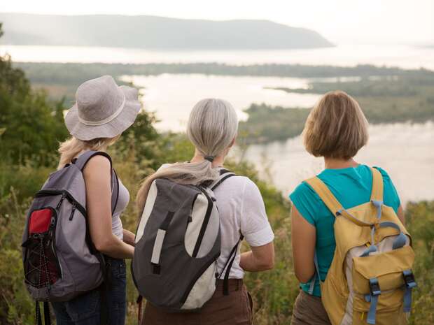 Three people with backpacks stand on a grassy hill, gazing at a serene lake and distant mountains under a clear sky.