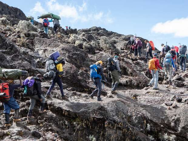 Hikers climb a rocky hillside, carrying large backpacks. They navigate the jagged terrain surrounded by sparse vegetation under a bright, partly cloudy sky.