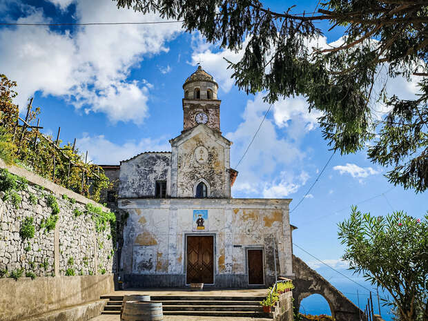 A rustic church with a clocktower stands quietly under a blue sky, surrounded by stone walls and greenery, overlooking a distant sea.
