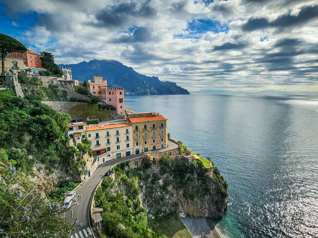 Coastal buildings with terracotta roofs sit atop a cliff, overlooking a calm sea. A winding road traces the cliff's edge, while distant mountains rise under a partly cloudy sky.