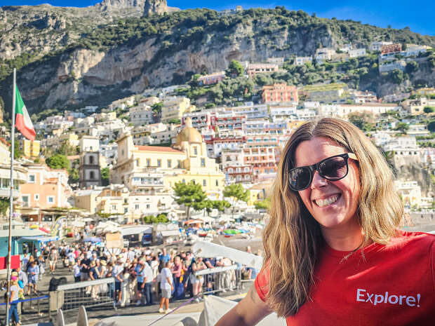 A smiling person wearing a "Explore!" shirt and sunglasses stands in front of a crowded town square with colorful hillside buildings and rugged cliffs in the background.