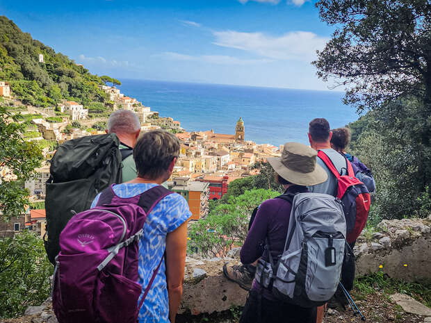 People with backpacks look out over a coastal village, surrounded by lush greenery and the sea in the background. They stand on a rocky path, admiring the view.