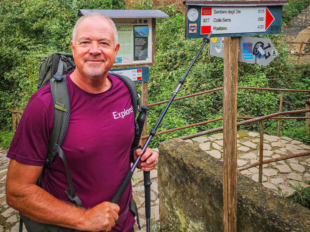 A man with a backpack and hiking pole stands beside a signpost showing trail directions: "Sentiero degli Dei 0:15," "Colle Serra 0:30," "Positano 4:20," against a lush, green backdrop.