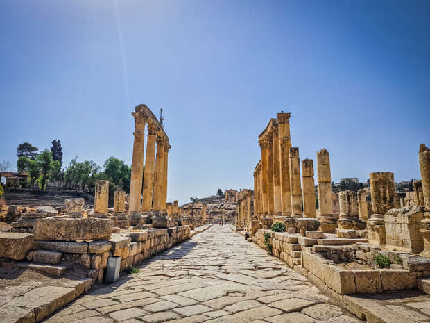 Ancient stone columns stand along a paved walkway, surrounded by ruins, under a clear blue sky. Greenery and distant hills are visible, indicating a historical archaeological site.