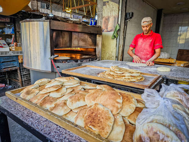 Baker shaping dough in a bustling bakery, with stacks of freshly baked flatbreads on trays. An open oven is visible behind, surrounded by kitchen utensils and equipment.